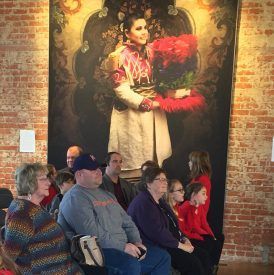 People seated, facing a large portrait of a woman in colorful attire, inside a brick-walled room.