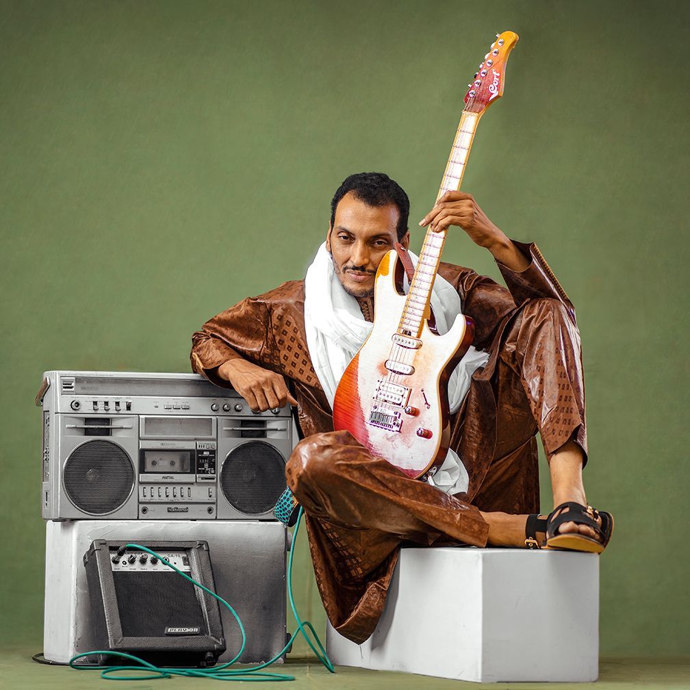 Man in brown outfit with guitar, white scarf, seated next to a boombox and amp on white blocks, green background.