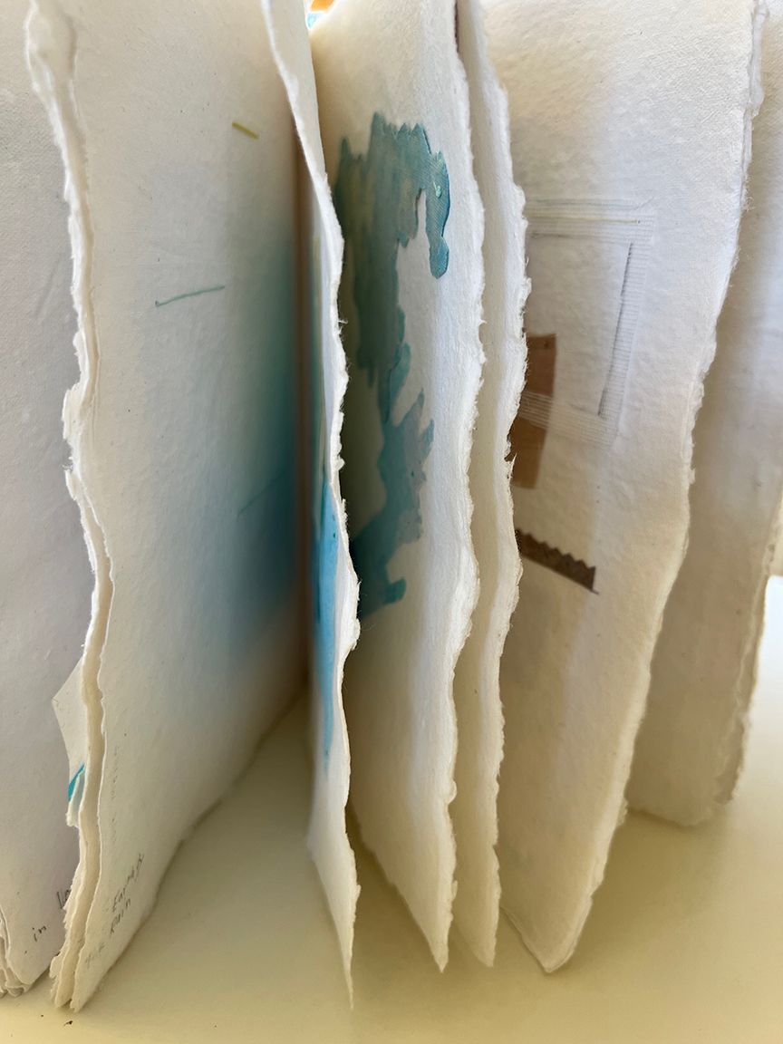 Close-up of a handmade book with rough, torn-edge pages. Some pages show watercolor art in shades of blue.