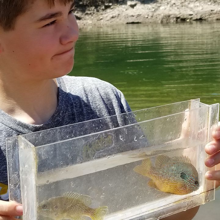 Boy holding a clear container with two fish in it, near a lake.