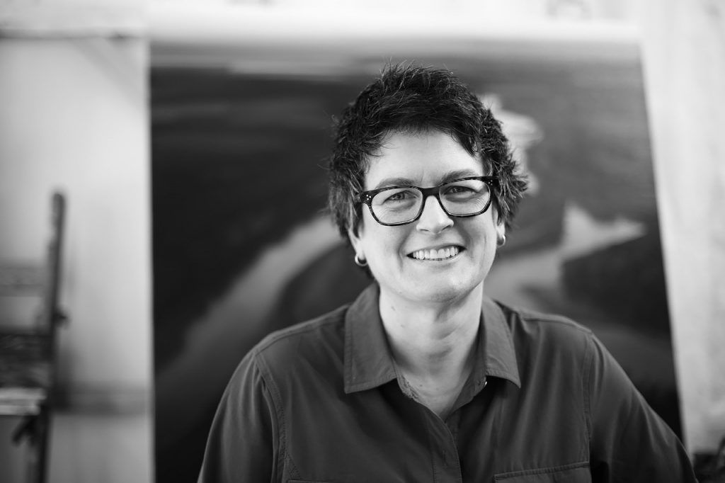 Woman with glasses smiles; black and white photograph in a studio with artwork in the background.