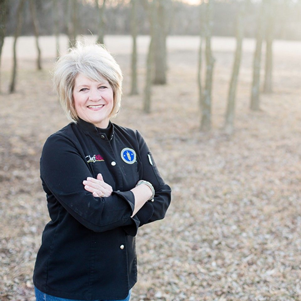 A woman is standing in a field with her arms crossed and smiling.