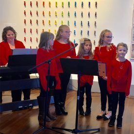 Choir of children in red sweaters sing in front of a keyboard player; colorful art on wall.