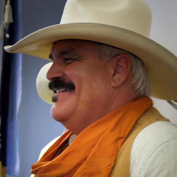 Man with mustache wearing cowboy hat, orange scarf, smiling.