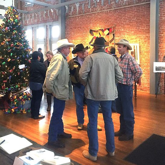Group of men in cowboy hats converse in a gallery with a Christmas tree and artwork on the walls.