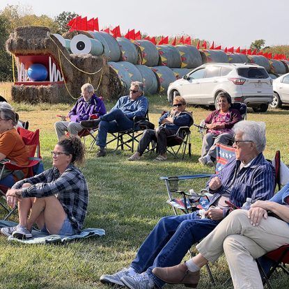 People seated in chairs watching an outdoor event next to a hay bale dragon sculpture.