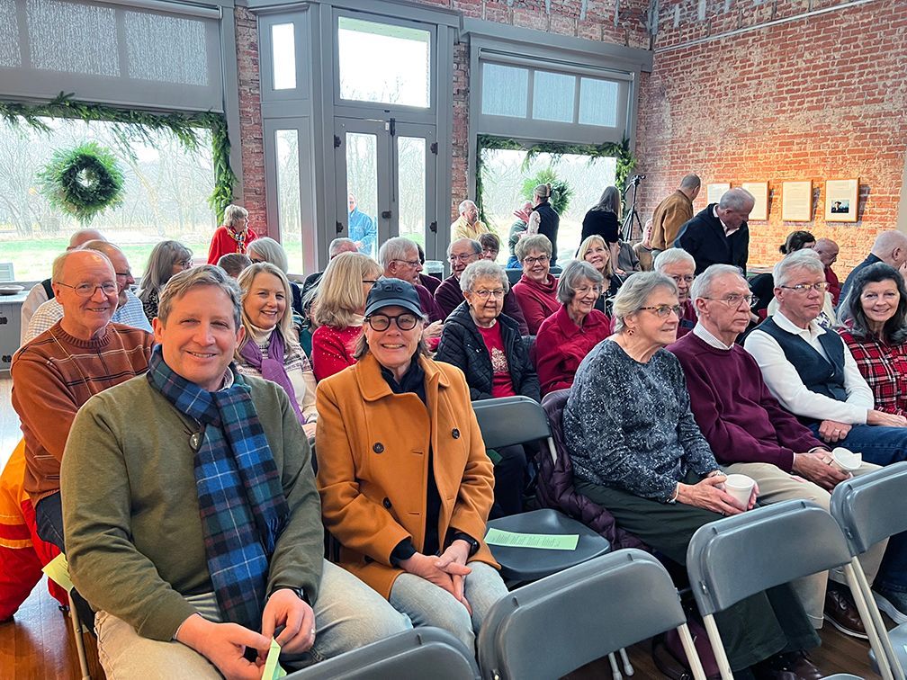 A seated crowd of smiling people facing the camera, with the view of the room's big store windows and a standing crowd of visiting audience behind them.