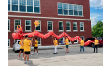 People in yellow shirts carry a red dragon in front of a brick building.