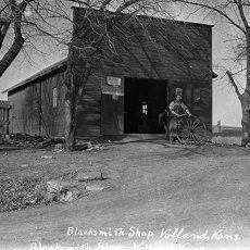 Black and white photo: Blacksmith shop in Volland, Kansas. Man with wheel outside open doorway.