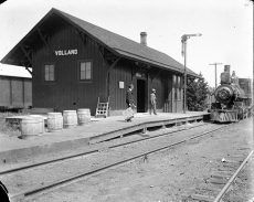 A black and white photo of the Villard train station with a train alongside the platform. Two people stand on the platform.