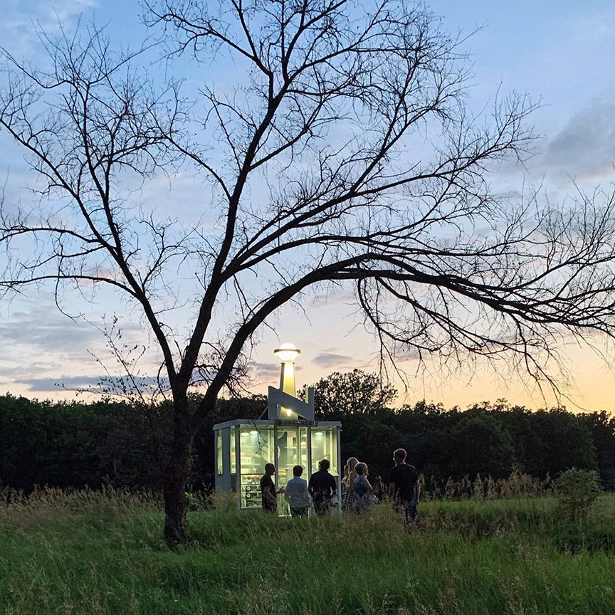 People gather in a glass structure under a bare tree at dusk in a field.