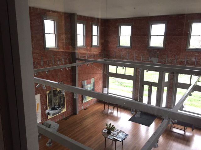 Interior view of a gallery with red brick walls, wooden floor, and large windows. Artworks hang on the walls.