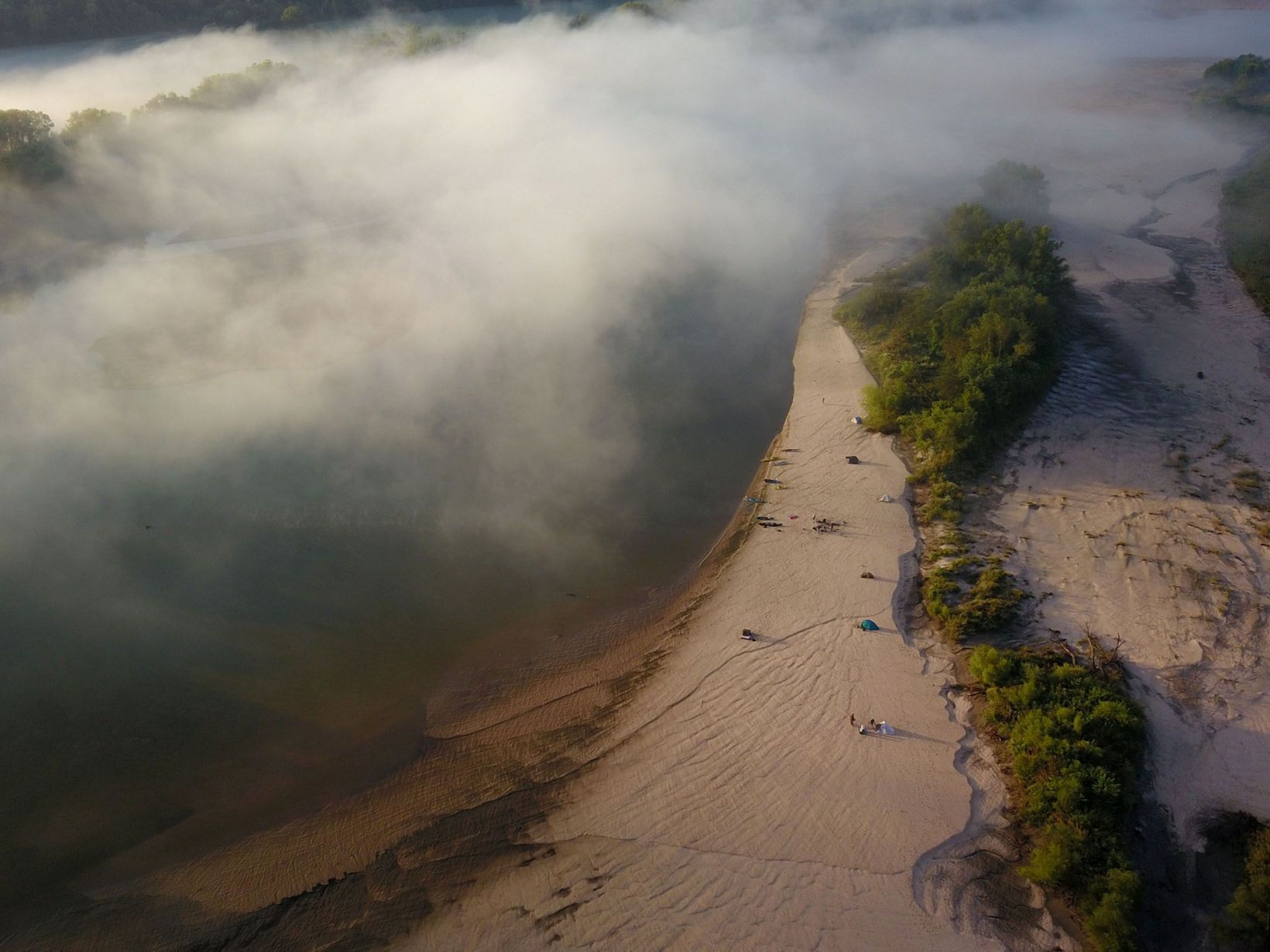 River landscape with low-hanging fog partially obscuring the water and sandy shore, with trees lining the shore.