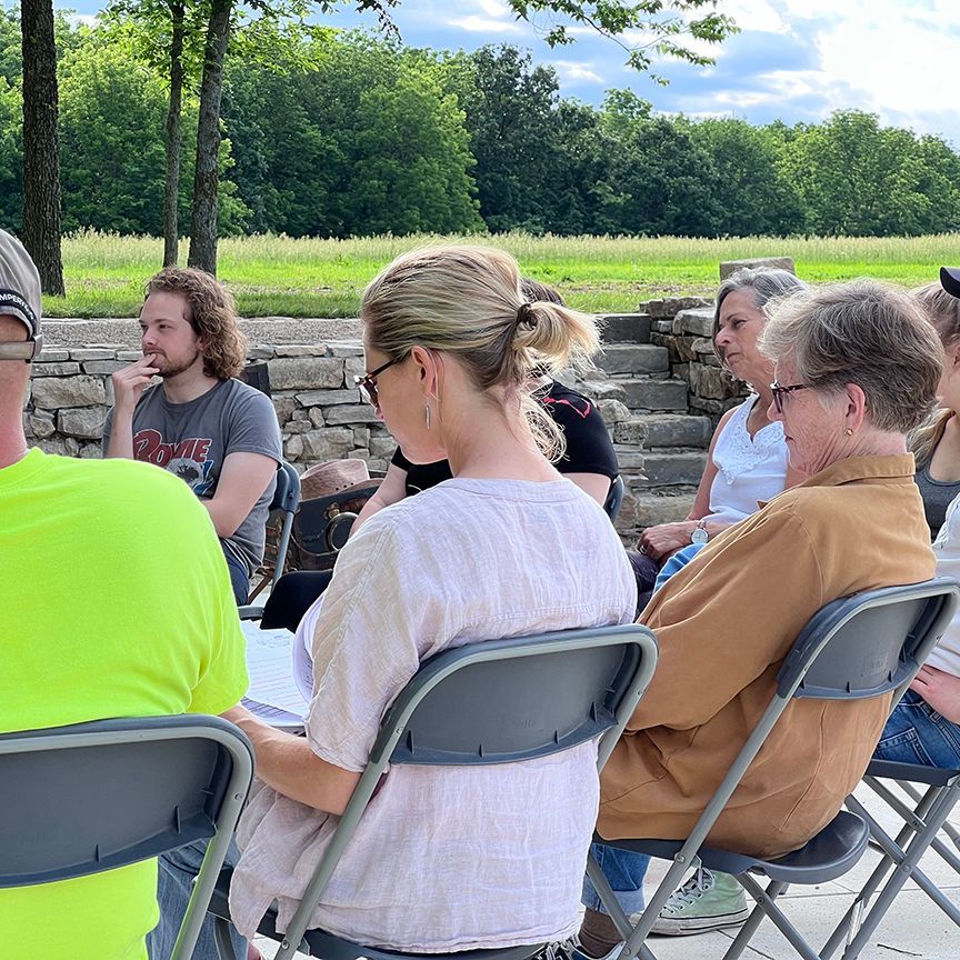 Group of people seated outdoors in chairs, listening. Light-colored hair, trees in background, sunny day.