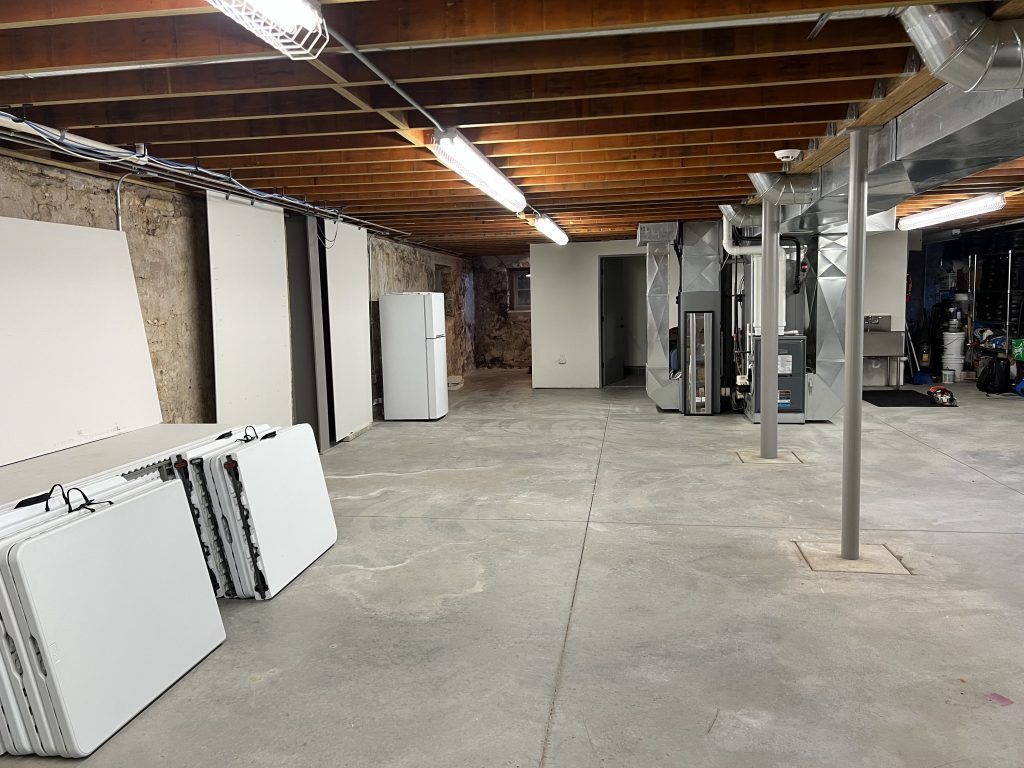 A dimly lit, concrete-floored basement with a refrigerator, stacked panels, and exposed beams and ducts.