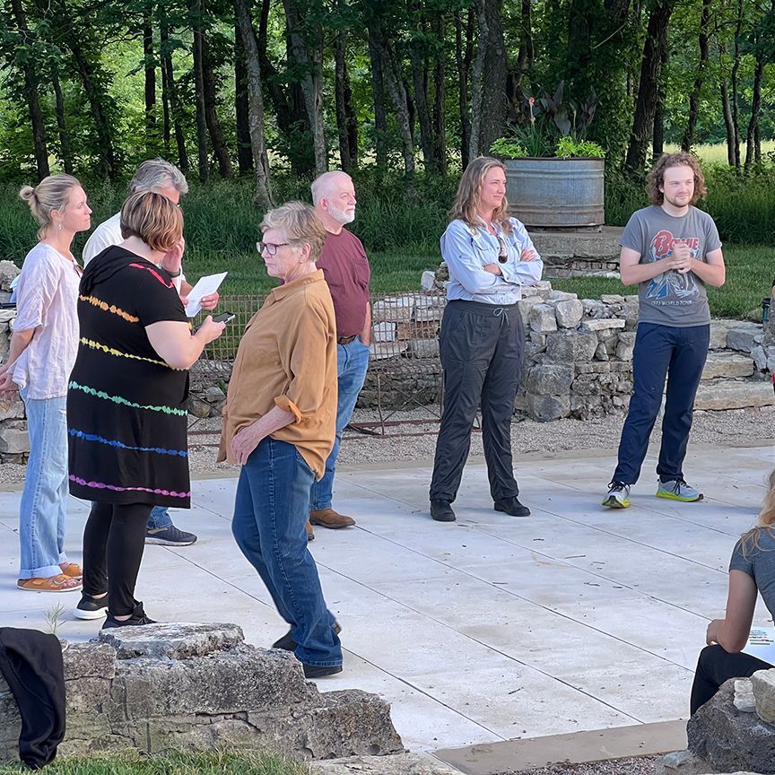 A group of people stand outside, some talking. Trees and stone ruins are in the background.