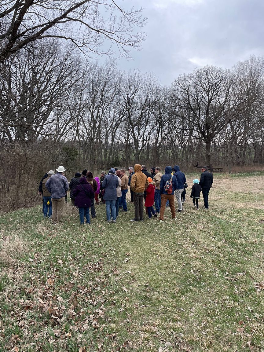 Group of people standing in a grassy field near bare trees, cloudy day.