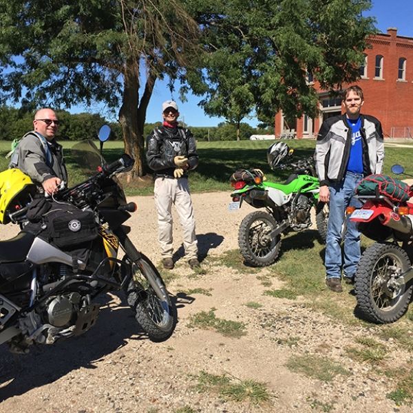 Three men are standing next to motorcycles in a dirt field
