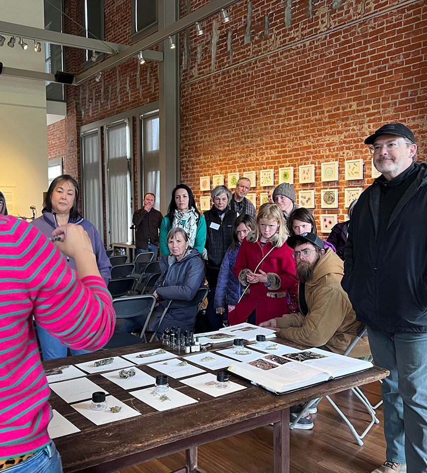 A group in an art gallery examines prints displayed on a table. Attendees of various ages and genders are gathered.