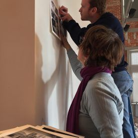 Two people hanging pictures on a wall; man on a step, woman looking up, both touching the pictures.