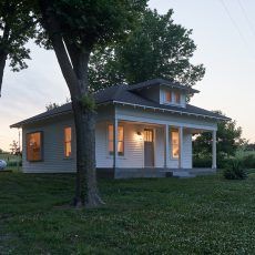 White house with porch, trees, and glowing windows at dusk.