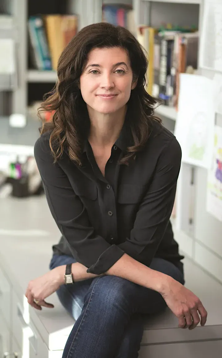 Woman in black shirt and jeans, sitting on a desk, smiling. Background is an office.
