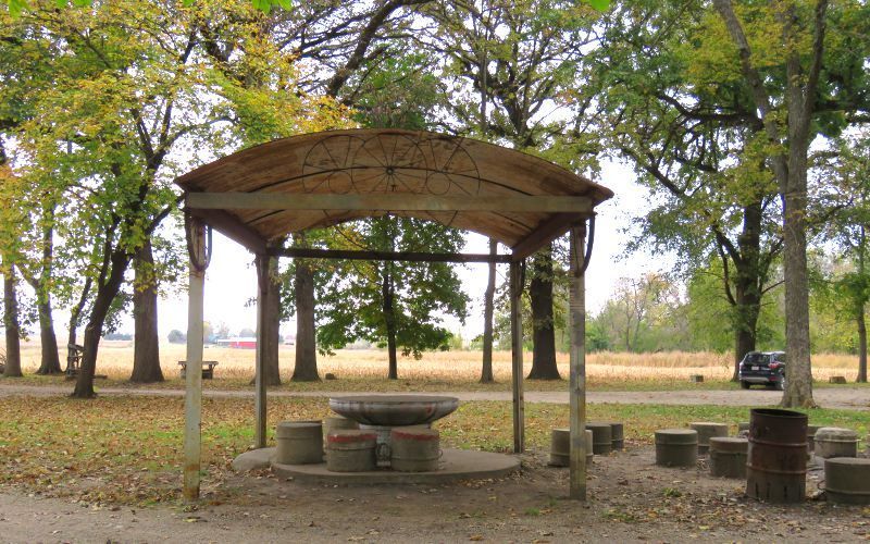 A rustic shelter with seating in a park, trees in the background, field in the distance.