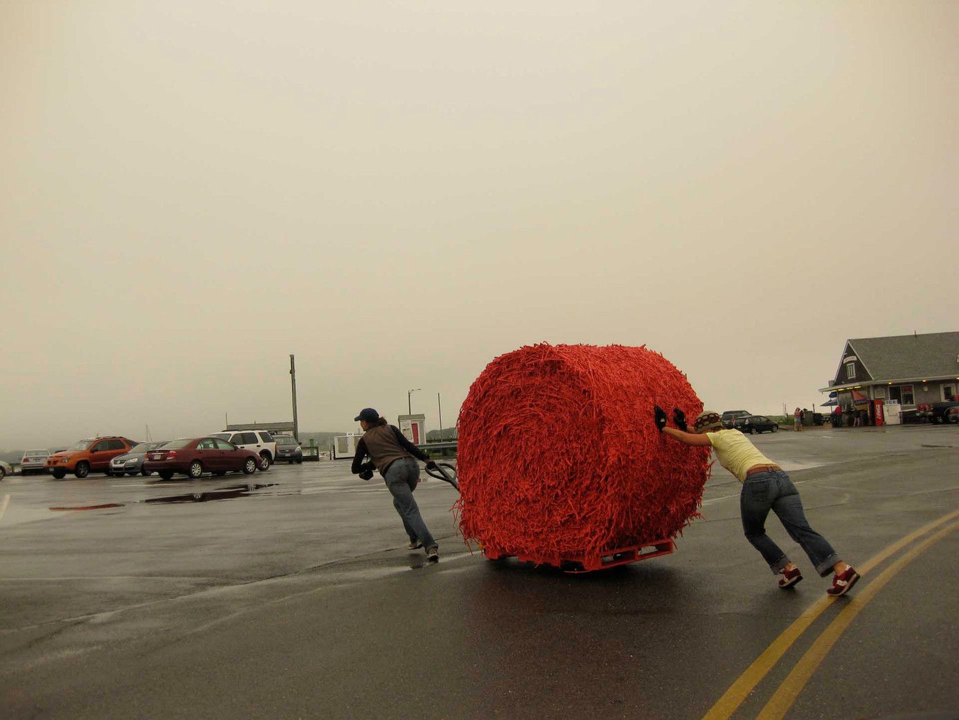 Two people on roller skates pull a large red sphere across wet pavement near parked cars.