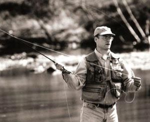 Man fly fishing in a river, holding a rod, wearing a vest and hat, with trees in the background.