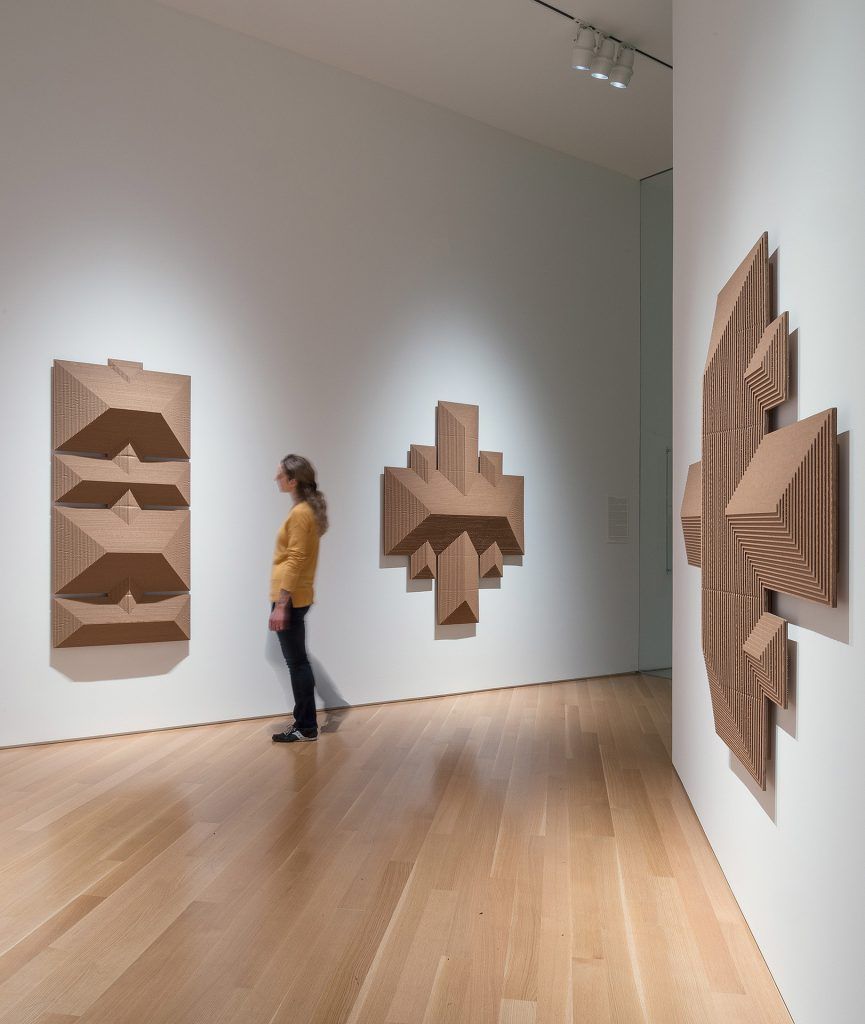 Woman viewing textured brown geometric sculptures in a gallery with hardwood floors and spotlights.