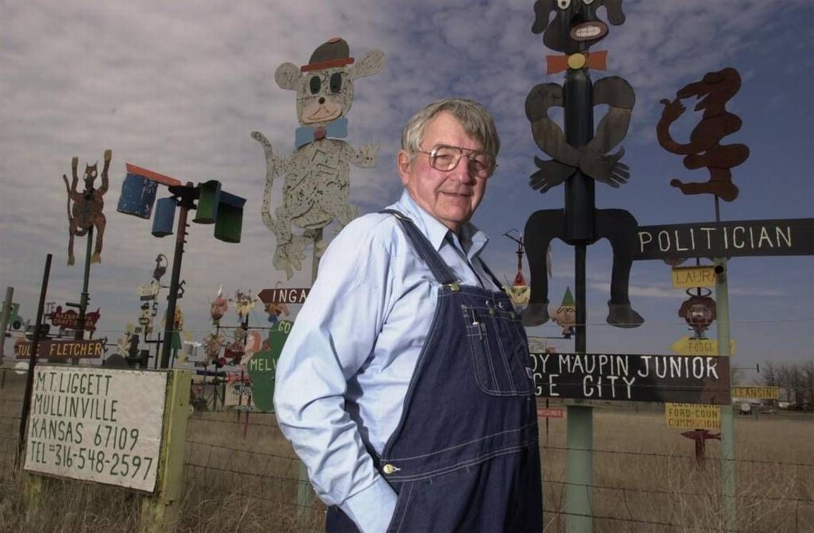 Man in overalls stands near quirky roadside folk art sculptures, including a dark-skinned figure.