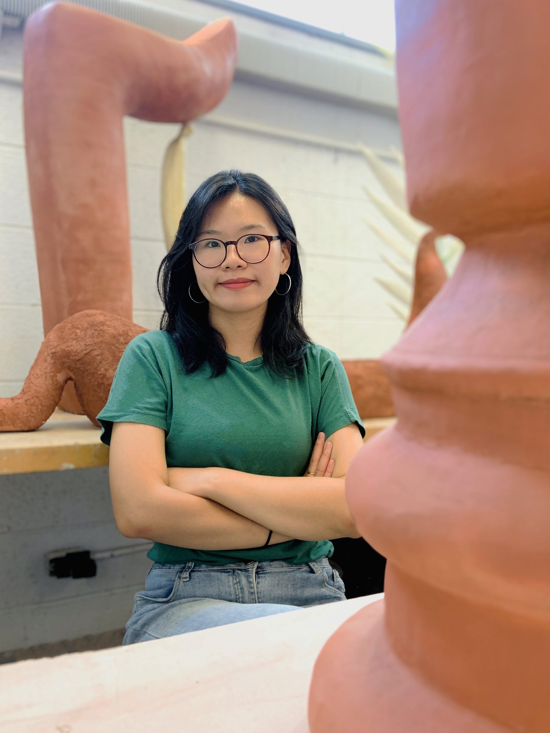 Woman in glasses with arms crossed, smiling in a pottery studio.