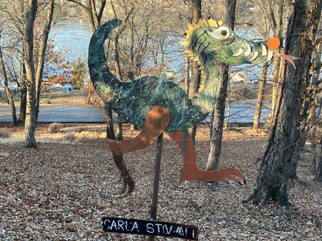 Whimsical, green and brown dragon sculpture in a yard, with a lake in the background.