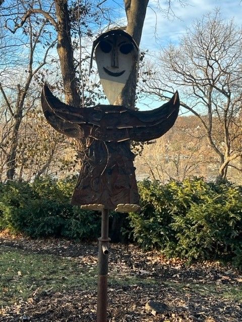 Rustic metal sculpture of a smiling face, wing-like arms, and a skirt in a park setting.
