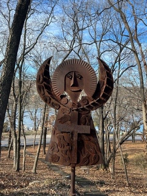 Rusty metal angel sculpture in a wooded area with a cross on its body and circular halo.