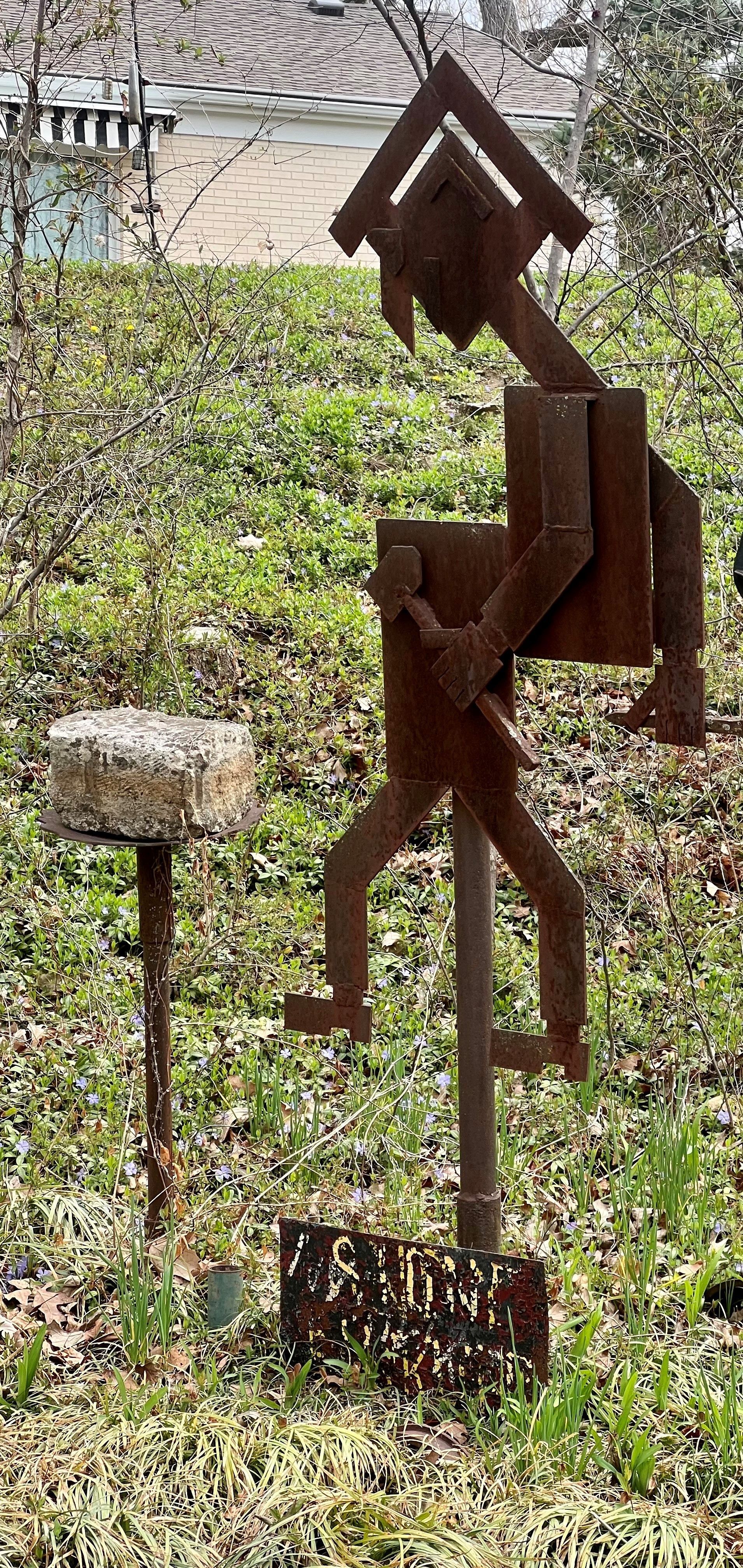 Rusty metal sculpture of a person with a house-shaped head, walking on stilts, in a grassy area.