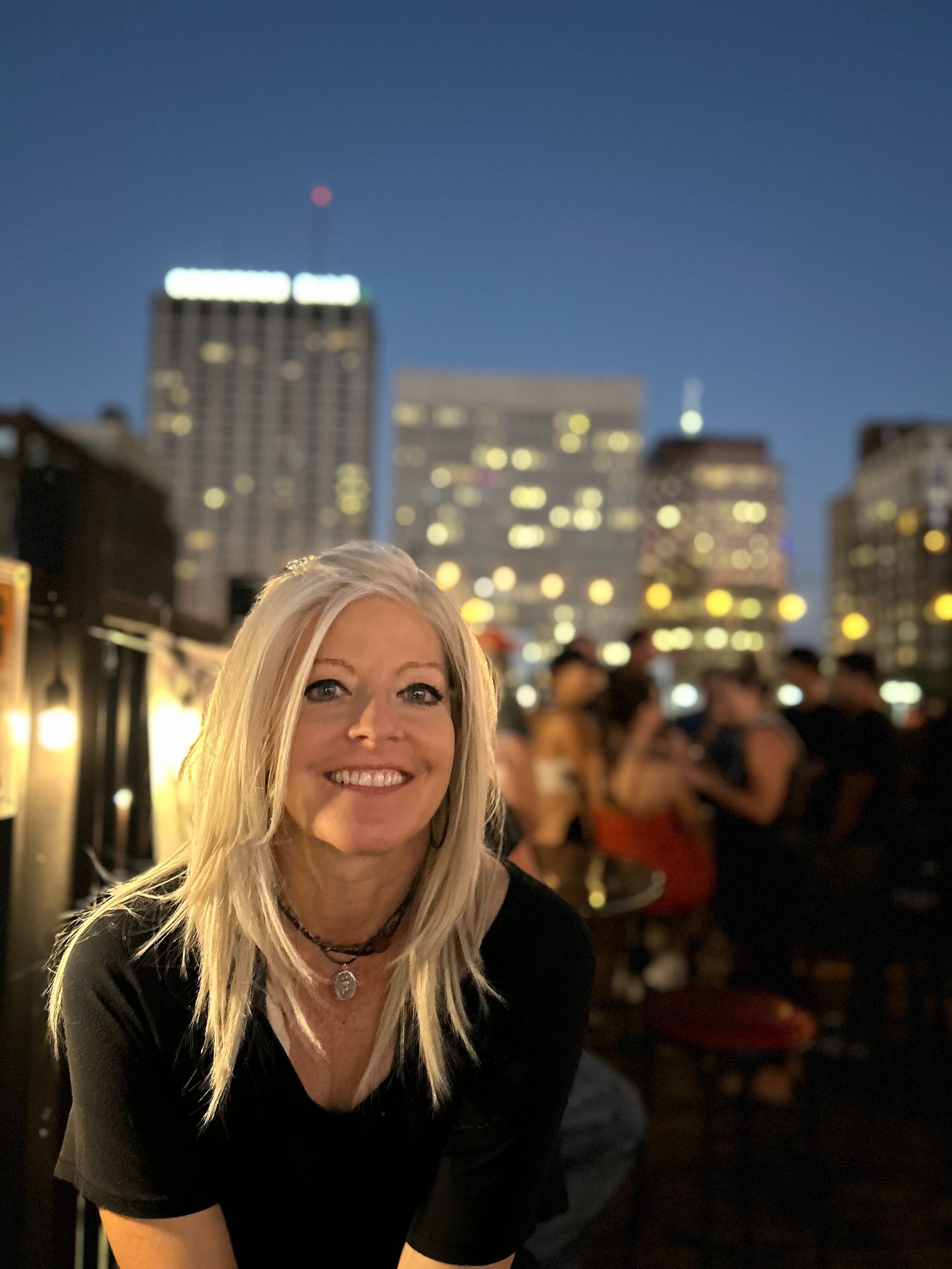 Woman with blonde hair smiling in front of a brick wall, wearing a green shirt and a crochet sweater.