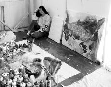 Artist smiles in studio, surrounded by paint supplies and canvases, one finished. Black and white.