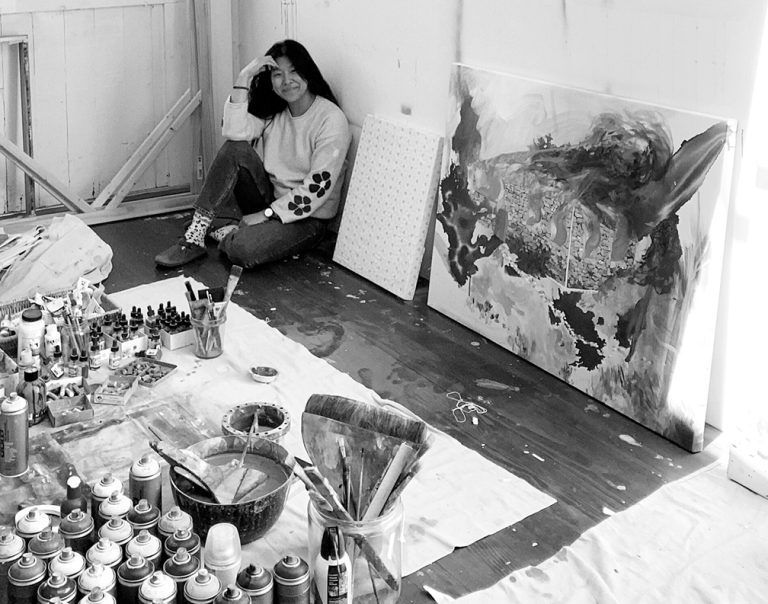 Artist sits in her studio, smiling. Paint supplies and abstract art surround her.