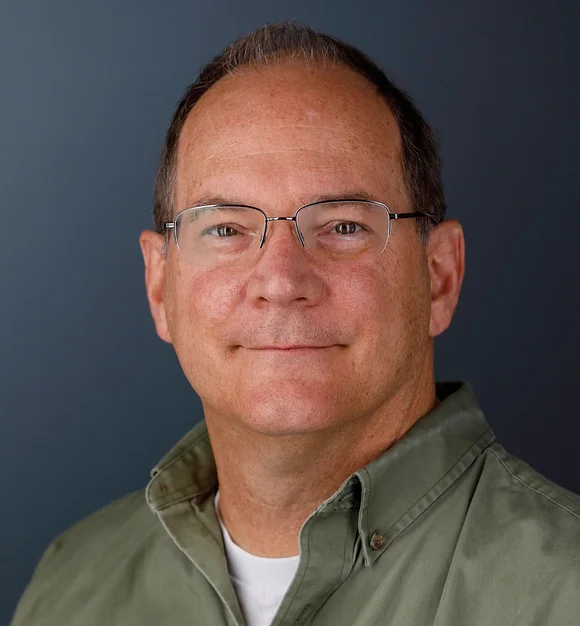 Man with glasses and a button-down shirt smiles, standing against a dark blue background.
