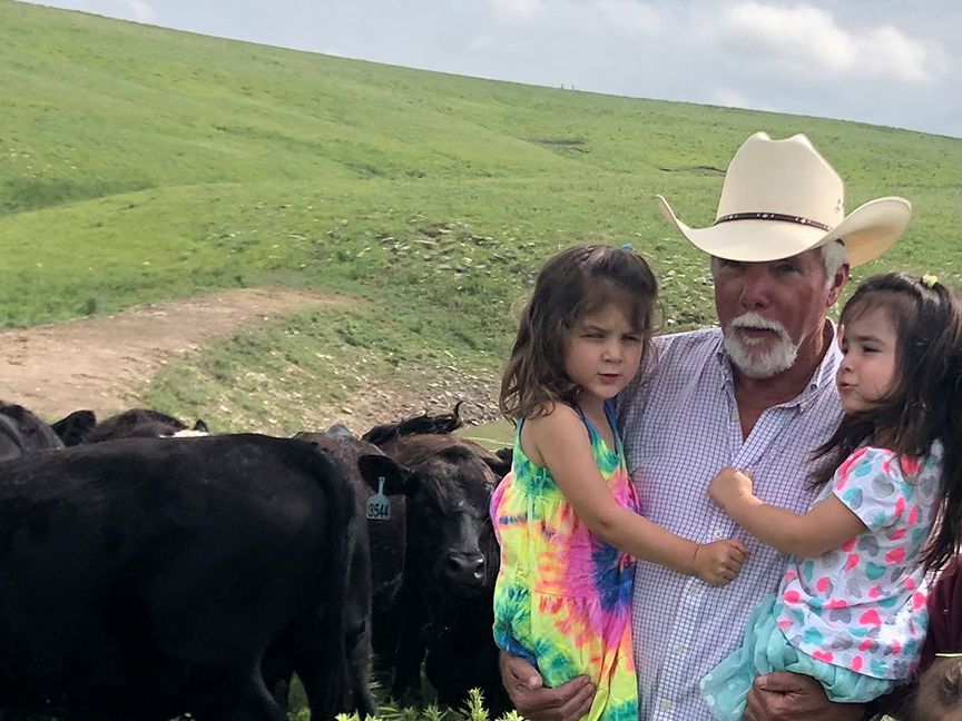A man in a cowboy hat is holding two little girls in front of a herd of cows.