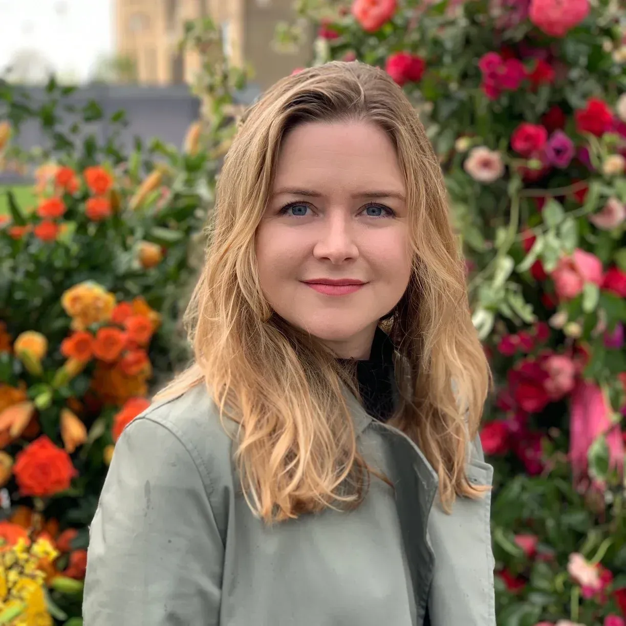 Woman with blonde hair smiles, standing in front of colorful flowers, wearing a green coat.