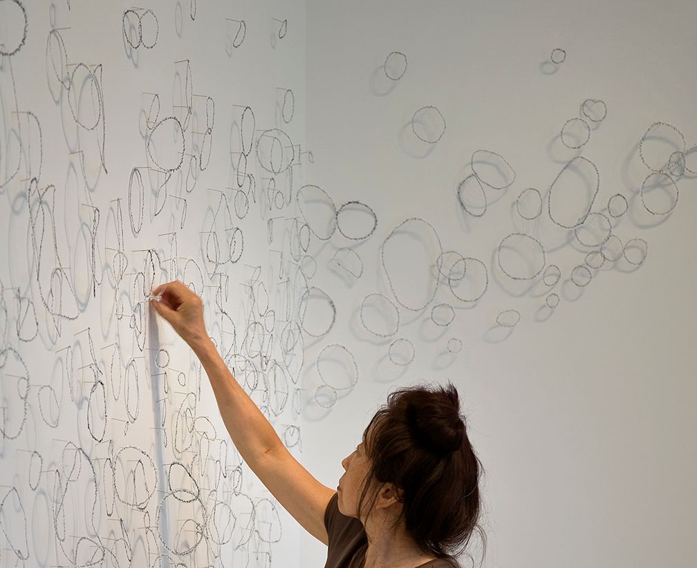 Woman draws on a wall with fine lines; many circles and lines are already drawn in a white room.