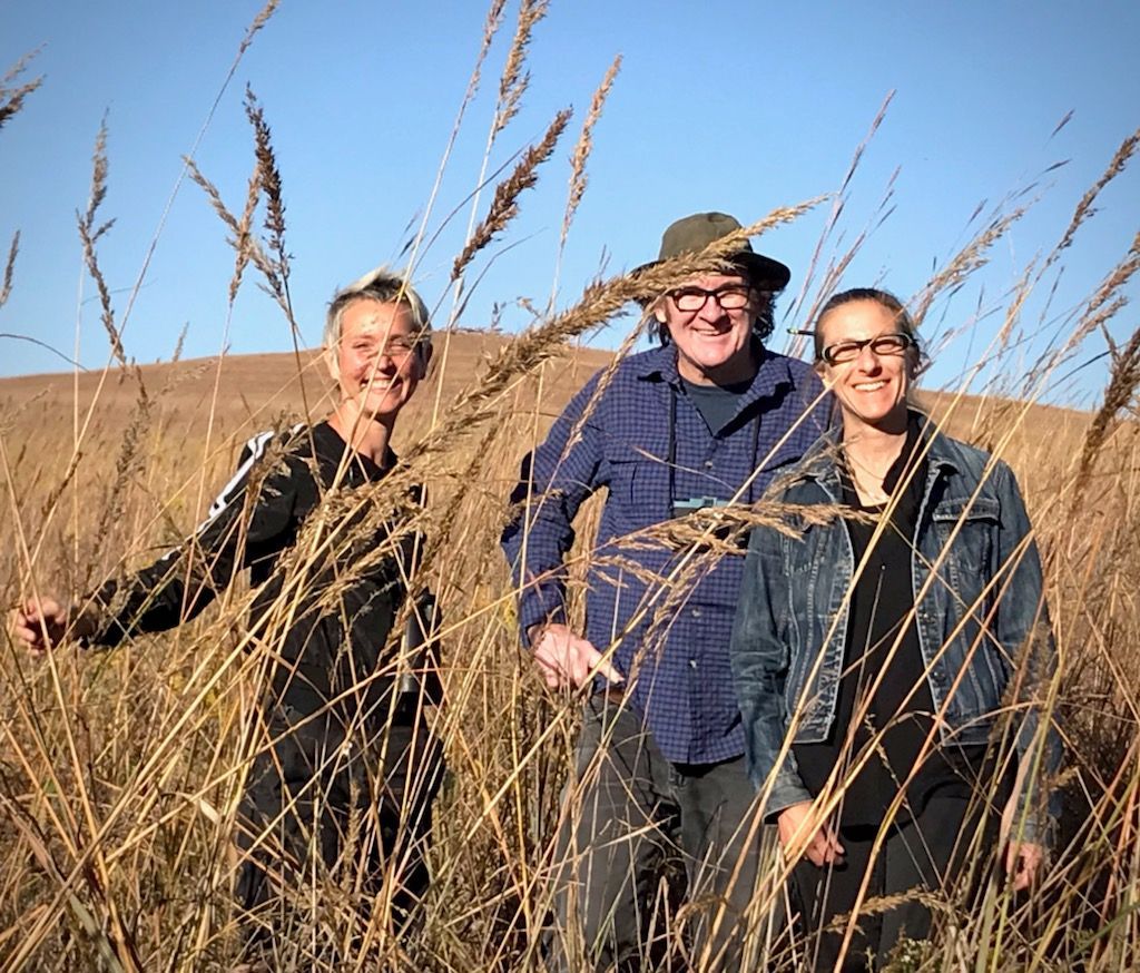Three people smiling in a tall, dry grass field under a blue sky.