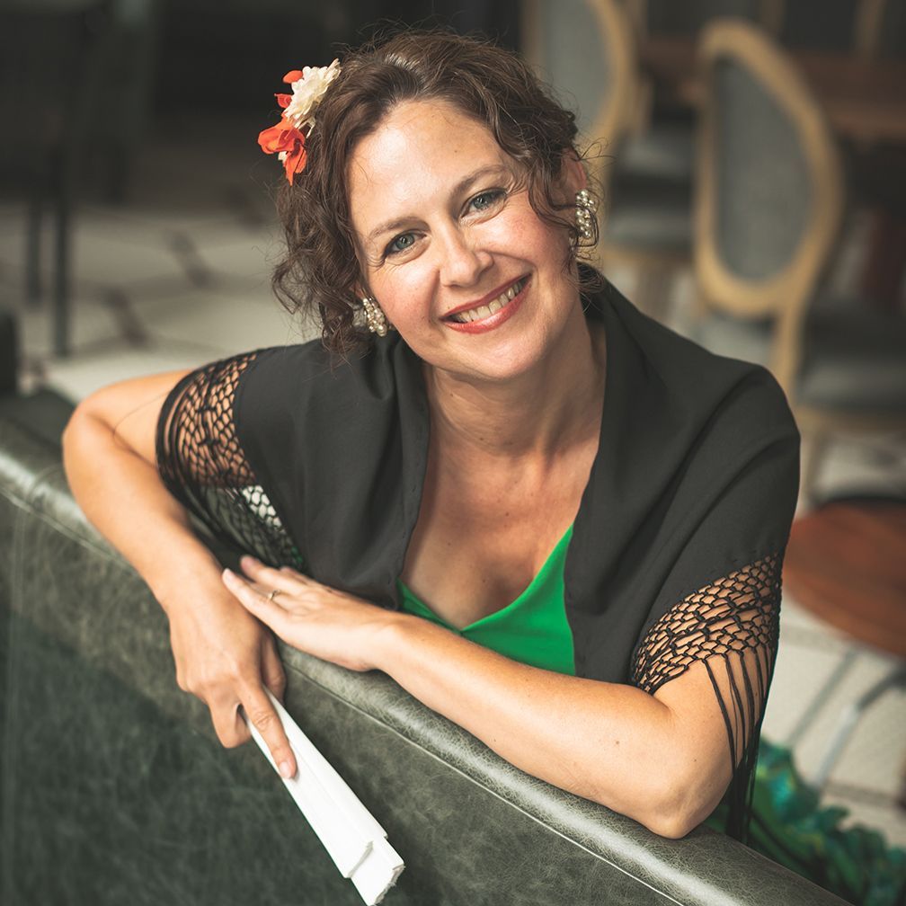 Woman with flower in hair, smiling, draped in black shawl, leaning on couch.