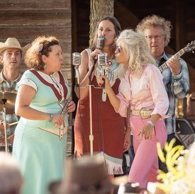 A band performs on an outdoor stage: women sing with microphones, musicians play instruments.