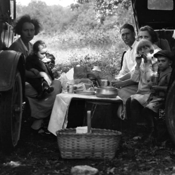 Family picnicking near a car in a wooded area; table set with food, basket in foreground, vintage.