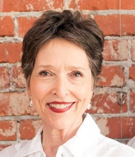 Woman with short brown hair smiles, red lipstick, white shirt, brick background.