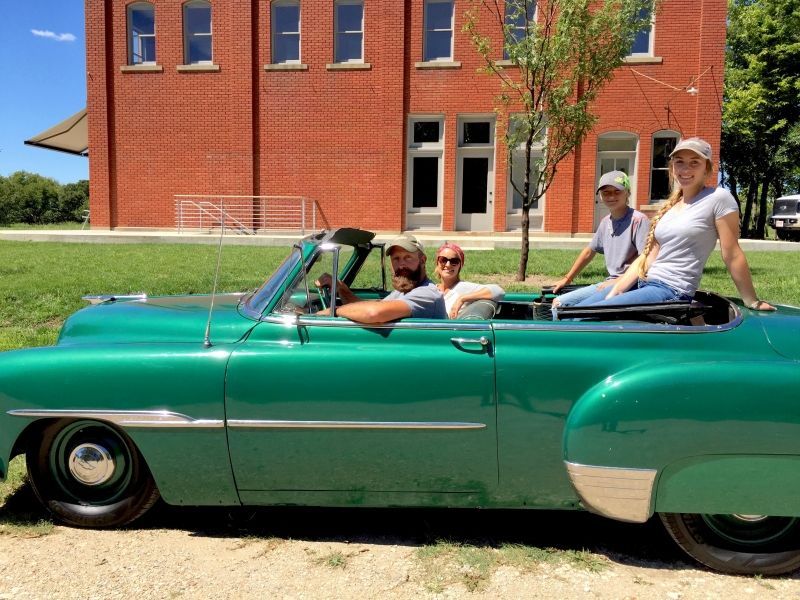 A group of people are sitting in a green convertible car in front of a brick building.
