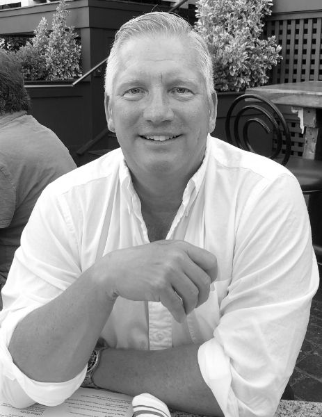 A man is sitting at a table with his hands folded and smiling in a black and white photo.
